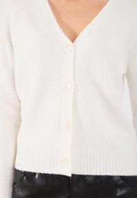 Biela cardigan s gombíkmi vyrobená z mäkkého materiálu. Má výstrih do V, rebrovaný lem a šesť krémových gombíkov na prednej strane.