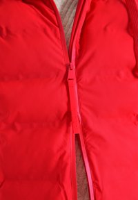 Rote Pufferweste mit einer glatten, glänzenden Außenseite, ausgestattet mit einem durchgehenden Reißverschluss und einem weichen, strukturierten Innenfutter.