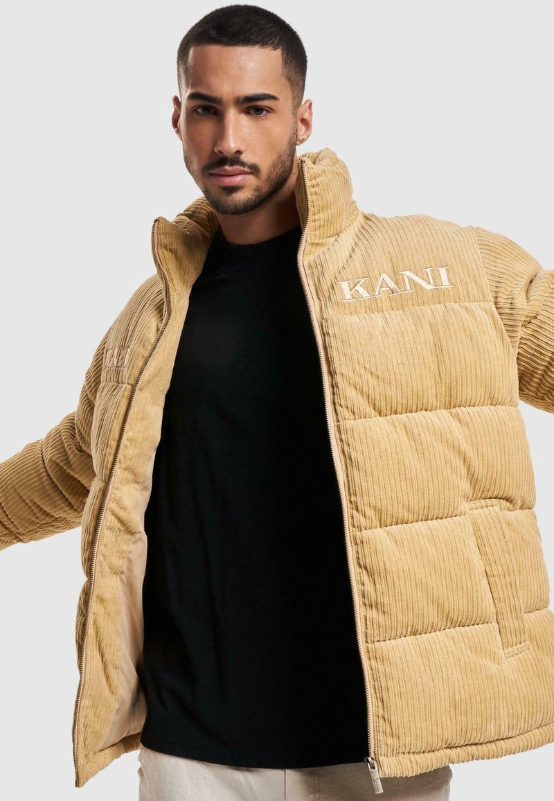 Chaqueta de plumas de pana color marrón claro con textura acolchada, cuello alto y logo bordado "KANI" en el pecho. Ligera, con cierre de cremallera y bolsillos laterales.
