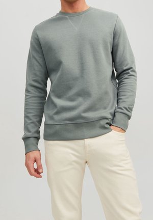 Homme portant un sweat-shirt gris à manches longues avec col rond et un pantalon blanc cassé, une main dans la poche, sur un fond clair uni.