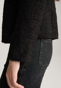 Zwarte, gestructureerde jas met een boxy pasvorm en kortere mouwen, gecombineerd met hoge zwarte denim jeans. Subtiel stofdetail zichtbaar.