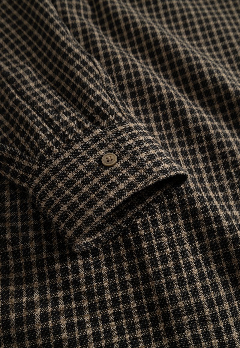 Tessuto a quadri nero e beige con una superficie texture. Presenta un bottone al polsino e un dettaglio di manica piegata.