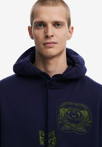 Marineblauwe hoodie met een trekkoord, voorzien van groene grafische ontwerpen aan de voorkant. Hete zachte textuur, casual pasvorm en vaste capuchon.