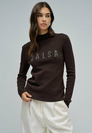 Suéter marrón de manga larga con un alto cuello y "SALSA" en brillantes piedras preciosas en la parte delantera, combinado con pantalones de color crema.
