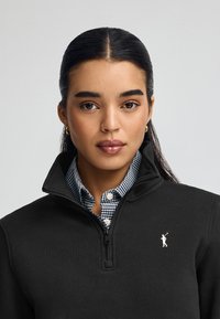 Suéter de forro polar negro con cremallera y alto cuello, adornado con un acento de logo blanco. Lleva una camisa de cuadros azul y blanco debajo. Textura suave.