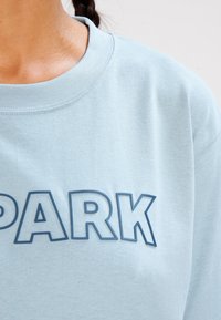 Sudadera de cuello redondo azul claro con una textura suave, que presenta un texto en negrita "PARK" en una impresión azul más oscura en el pecho.