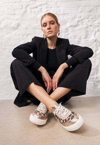 Leopardenmuster Sneakers mit goldenen Akzenten, weißen Sohlen und dickem Plateau, präsentiert mit einem schwarzen Anzug und neutralem Hintergrund.