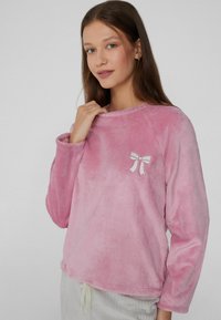 Top de forro polar rosa de manga larga con cuello redondo y un bordado de un lazo blanco en el lado izquierdo. Textura suave y ajuste relajado.