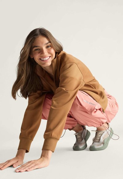 Brauner Langarm-Sweatshirt, rosa weite Hose und grau-grüne Sneakers. Das Outfit zeichnet sich durch entspannte Passformen mit sichtbaren Nähten und Texturdetails aus.