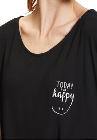 Svart kortärmad topp med rund halsringning och ett vitt tryckt motiv som säger "IDAG ÄR JAG glad" med en leende ansikte.