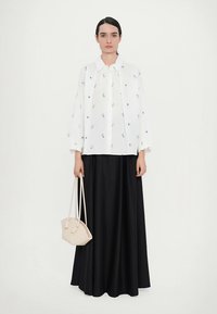 Vit skjorta med grön blomsterbroderi, kombinerad med en svart, flödande maxikjol. Håller en liten beige handväska med vävd textur.