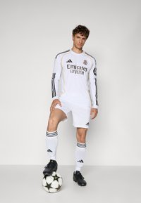 Maglia da calcio bianca a maniche lunghe con dettagli neri e dorati, con il marchio "Emirates", abbinata a pantaloni corti bianchi e scarpini neri, in piedi su un pallone da calcio.