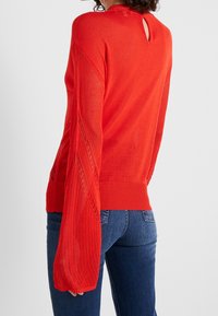 Red knitted sweater featuring long bell sleeves, openwork pattern on sleeves, and a back keyhole detail. Ribbed hem and cuffs.