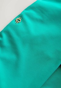 Tkanina w turkusowym kolorze o gładkiej fakturze, z dołączonym z boku złotym okrągłym emblematem z literą 'H'.