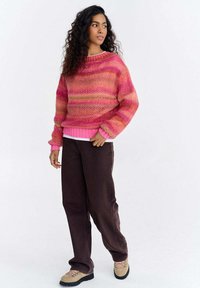 Kobieta o kręconych włosach, ubrana w różowo-pomarańczowy sweter w paski, ciemnobrązowe spodnie i beżowe buty, stojąca na białym tle.