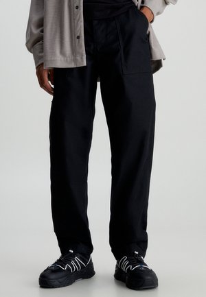 Man draagt zwarte losse broek, zwarte sneakers met witte veters en een beige jasje met knopen, met één hand in de zak.