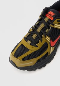 Close-up de um sneaker preto e oliva da Nike com logótipo vermelho e cordões pretos sobre fundo branco.