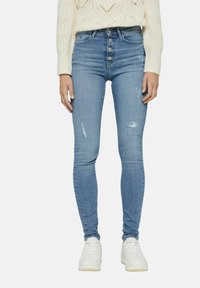 Jeans ajustados de color azul claro con una cintura alta, cierre de cinco botones y detalles desgastados en las rodillas. Combinados con zapatillas blancas.