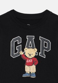 Čierne tričko s veľkým nápisom "GAP" obrysom v bielej farbe a kresleným medveďom oblečeným v červenom svetri GAP, béžových šortkách a modrých topánkach, ktorý máva jednou rukou.