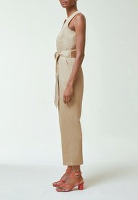 Femme en combinaison beige sans manches avec ceinture à la taille, portant des sandales corail à talons blocs et à brides, debout de profil sur un fond clair uni.