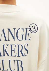 Crème sweatshirt met grote marineblauwe tekst die "CHANGE MAKERS CLUB" leest, met een kleine geborduurde glimlach op de schouder.