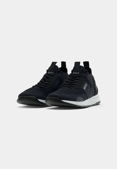 Baskets de sport noires avec une tige en tissu texturé, semelle blanche contrastante et détail du logo sur la languette. Présente un design à lacets et une languette de traction.