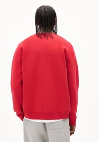 Felpa rossa a girocollo realizzata in un tessuto morbido, con una vestibilità comoda e polsini a costine. Indossata sopra pantaloni della tuta grigi.