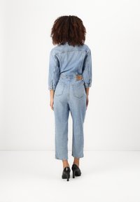 Denim kombinezon svetlo modre barve, dolgih rokavov, z zipperjem v pasu in ravnimi hlačami, z zadnjimi žepi in blagovno oznako v rjavi barvi.