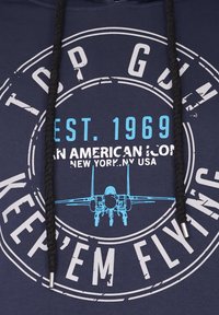 Mörkblå hoodie med en grafisk design som har vit och blå text. Texten inkluderar "TOP GUN", "EST. 1969" och en illustration av ett jetplan. Svarta dragsnören.