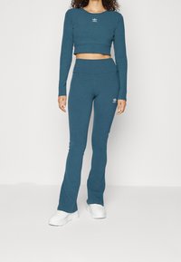 Žena v námořnickém modrém upnutém dlouhém rukávu a odpovídajících rozšířených legínách se bílými teniskami, stojící na bílém pozadí.