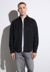 Camisa de pana negra con cuello abotonado, bolsillo en el pecho, mangas largas y dobladillo redondeado, llevada sobre una camiseta blanca.