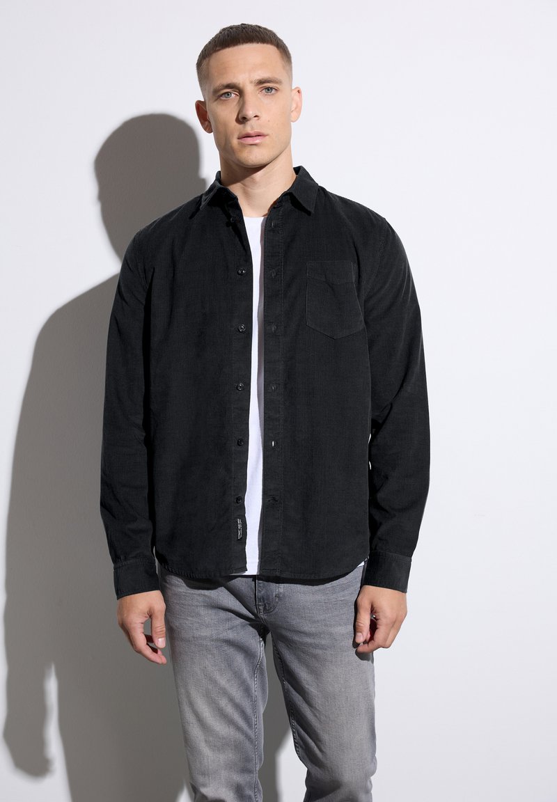 Camisa de pana negra con cuello abotonado, bolsillo en el pecho, mangas largas y dobladillo redondeado, llevada sobre una camiseta blanca.