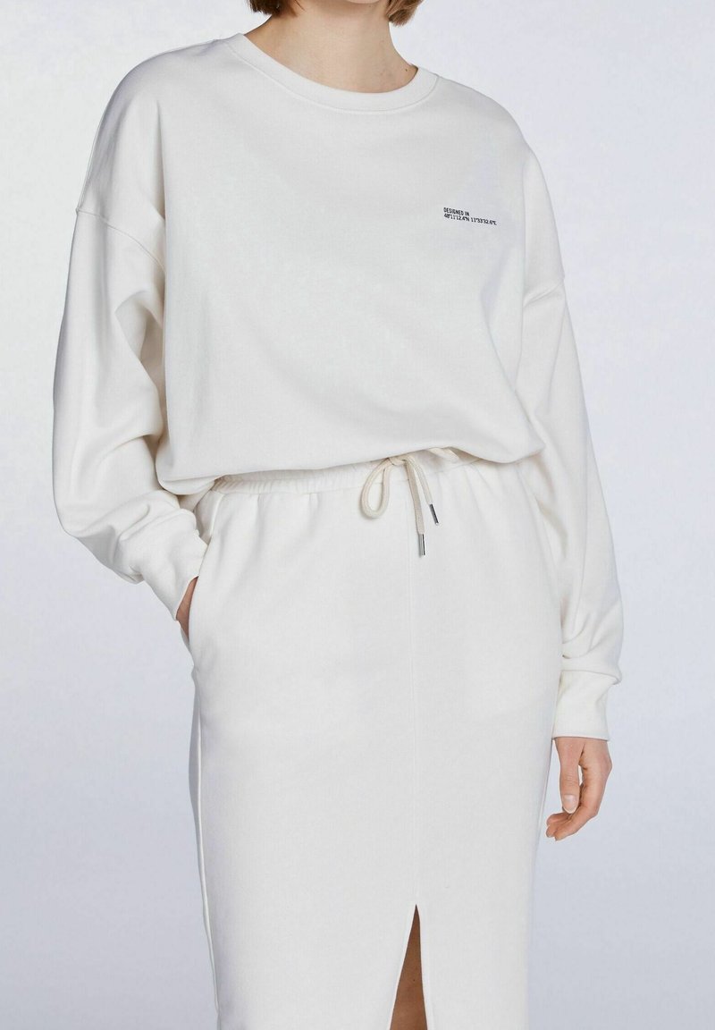 Conjunto de sudadera y falda blanca, hecho de una tela suave. La sudadera presenta un escote redondo y mangas largas, mientras que la falda tiene una cintura con cordón y una abertura lateral.