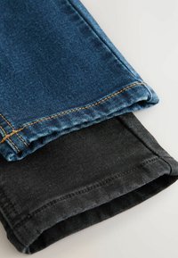 Două perechi de blugi cu manșete: una din denim albastru cu cusături aurii, cealaltă din țesătură de bumbac negru, aranjate una peste alta pe un fundal deschis.