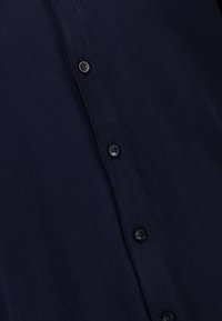 Marinblå cardigan med strukturerat stickat, som har en rad svarta knappar framtill och en klassisk kragdesign.