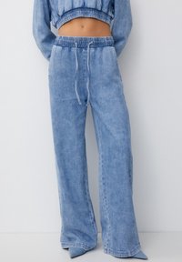 Los pantalones de mezclilla anchos en un suave tono azul claro cuentan con una cintura elástica con cordón y bolsillos laterales. La tela tiene un acabado suave y ligeramente texturizado.