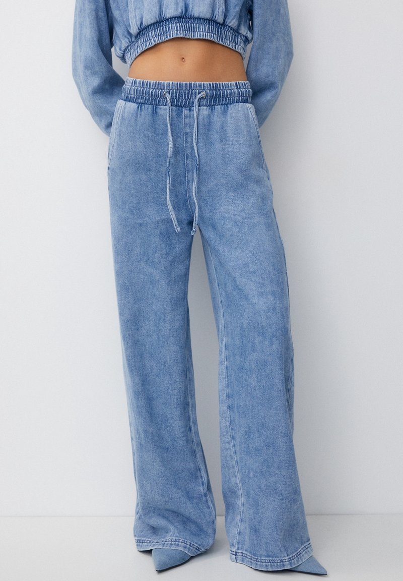 Los pantalones de mezclilla anchos en un suave tono azul claro cuentan con una cintura elástica con cordón y bolsillos laterales. La tela tiene un acabado suave y ligeramente texturizado.