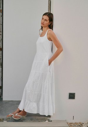 Nő ujjatlan, fehér maxiruha zsebekkel, egy világos rózsaszín falnak támaszkodva, fehér flip-flop papucsot viselve kőlépcsőn.