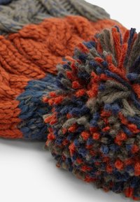 Gebreide beanie met een mix van oranje, blauw en grijze garens, textuurkabels en een grote, pluizige, meerkleurige pompon bovenop.