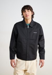Veste zippée noire avec un col classique et des poches latérales, arborant le logo blanc "Carhartt" sur la poitrine gauche. Fini en tissu lisse.