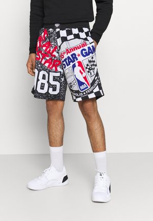 Persona che indossa pantaloncini NBA All Star Game neri e bianchi con testo rosso e blu, calzini bianchi e scarpe da ginnastica bianche, in piedi su pavimento grigio.