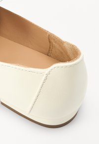 Vita läder slip-on sko med rundad baksida, mjukt mockainteriör och en tan gummisula. Sömdetaljer längs kanterna.