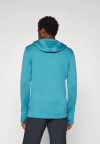 Moški v turkizni kapuci, dolgih rokavih in črnih hlačah, prikazan od zadaj na enobarvni svetli podlagi.