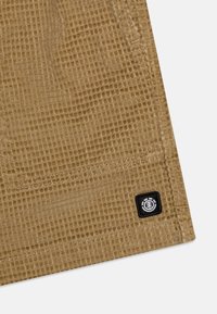 Tissu brun texturé avec un motif en grille, présentant une étiquette noire avec un logo dans le coin inférieur et des détails de couture visibles.