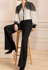 Svarta vida byxor i kombination med en vit blus med svarta prickar, dekorativa linjer och genomskinliga puffärmar med texturerade manschetter.