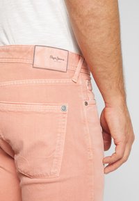 Jeans de mezclilla de color durazno con bolsillos traseros, detalles de metal y una etiqueta de cuero con marca. Las costuras están visibles.