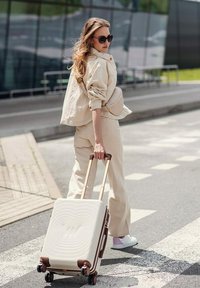 Tvrdý kufr v krémové barvě s texturovaným vlnkovým vzorem, bronzovými akcenty, čtyřmi kolečky a teleskopickou rukojetí. Model v béžovém outfitu.