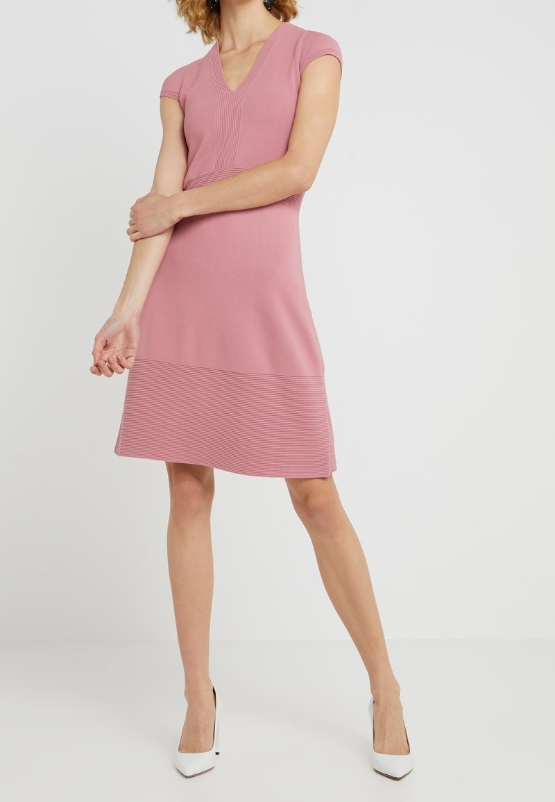 Rosa, knielanges Kleid mit kurzen Ärmeln, V-Ausschnitt, gerippter Textur am Oberteil und Saum, kombiniert mit weißen High-Heels.