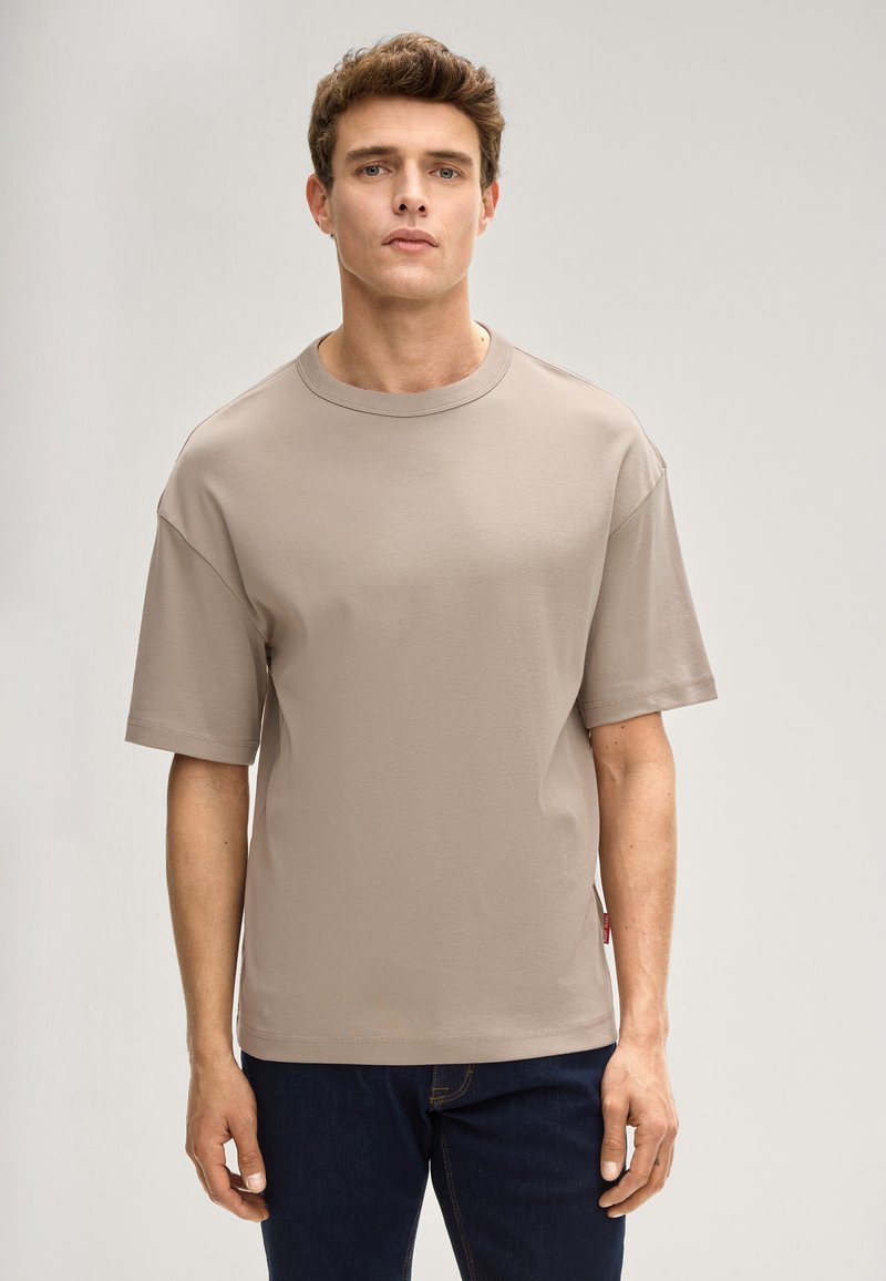 Beżowy t-shirt z krótkim rękawem wykonany z miękkiego materiału, z okrągłym dekoltem, luźnym fasonem i małą metką z logo po boku. Połączony z ciemnymi dżinsami.