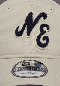 Căciulă bej, ajustabilă, cu un logo brodat „NE” de culoare bleumarin. Prezintă un material texturat și un eticheta rotundă cu inscripția „9TWENTY ADJUSTABLE.”